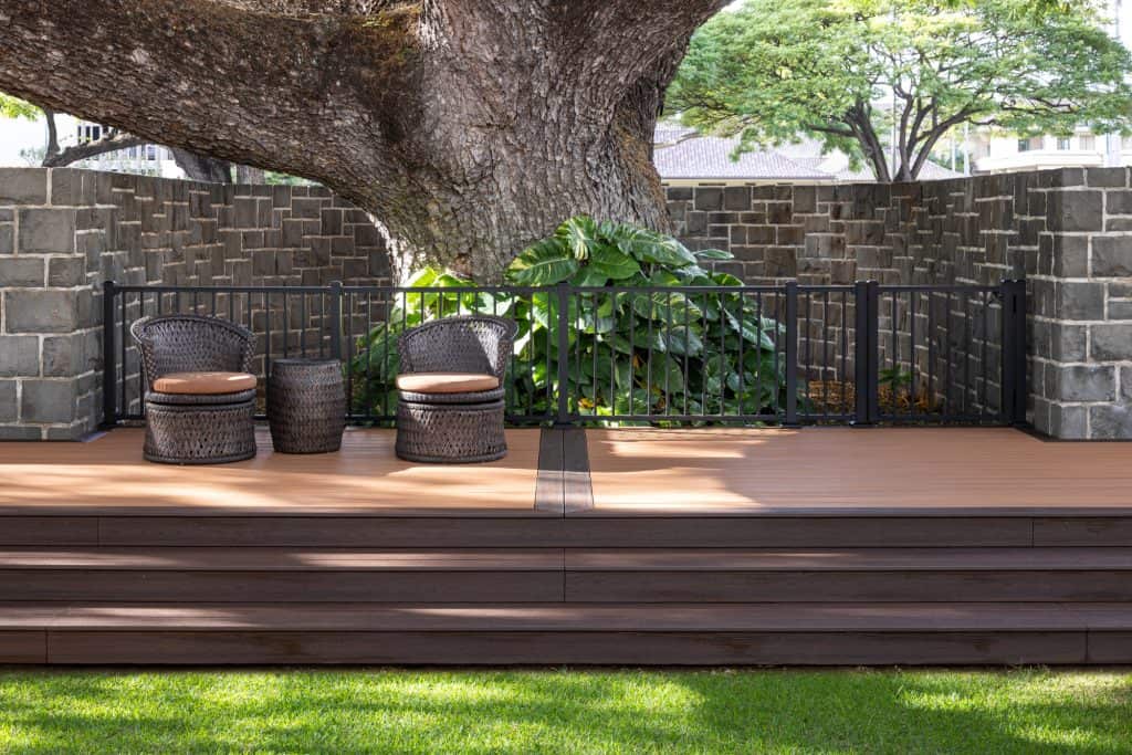 Front view of a deck built around a Hawaii State Exceptional Monkey Pod tree, showcasing the steps leading up to a Trex Transcend wedding platform. The Trex Black Signature railing and gate are installed in front of the tree for protection. The photo also features two wicker chairs with a small table, complementing the outdoor space. In the background, part of a brick wall surrounding the tree is visible, highlighting the integration of nature and craftsmanship in this Honolulu outdoor living space.