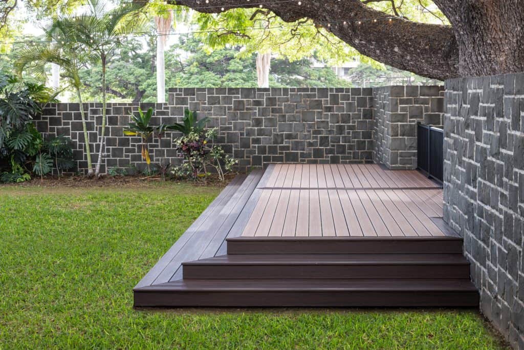 Side profile of a wedding platform built with Trex Transcend deck boards in Tiki Torch and Lava Rock, showcasing the craftsmanship of the steps leading up to the platform. The photo also features Trex Black Signature railing on the side and lush greenery, highlighting the contrast of the two colors selected for the deck and the precision in step construction. This modern outdoor space in Honolulu reflects quality design and craftsmanship.