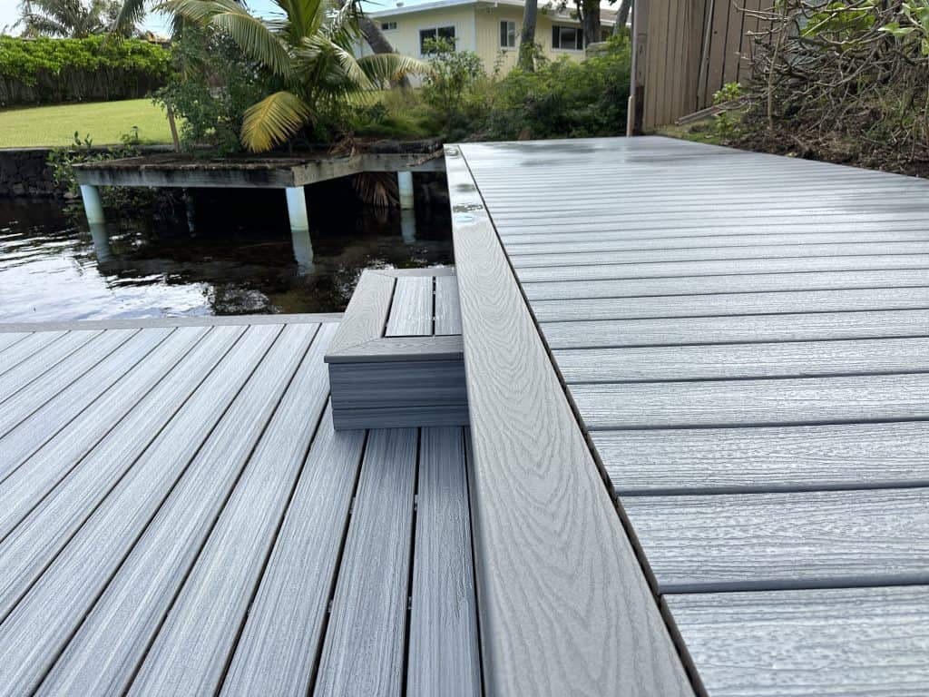 Side angle view of a two-level deck in Kailua, featuring Trex Foggy Wharf deck boards with Pebble Gray picture frame border. The top deck steps down to a lower level that overlooks a lake, showcasing exceptional craftsmanship in working near water. Lush greenery surrounds the area, enhancing the serene outdoor space.