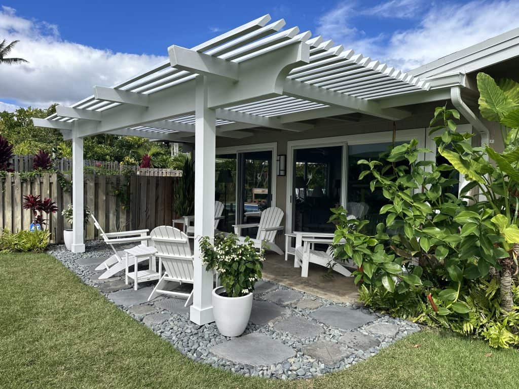 Wide-angle view of a custom white pergola with angled slats, complemented by new landscape pavers and outdoor seating in Kailua, Oahu. This outdoor living space offers sun protection, blending style and function in a tropical retreat. The pergola enhances the aesthetic and functionality of the space, providing an ideal environment for relaxation or entertaining guests.