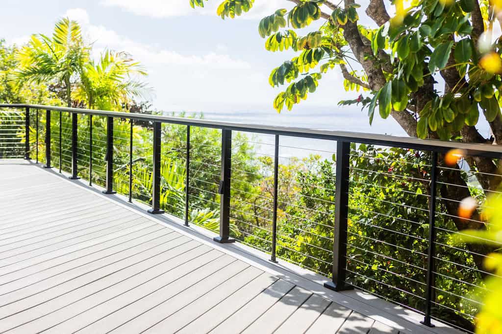 Side angle of the Azek TimberTech PVC deck boards in Vintage Coastline with Feeney stainless steel cable railing, showing off the double border and diagonal design. The background features tropical greenery and the Pacific Ocean.