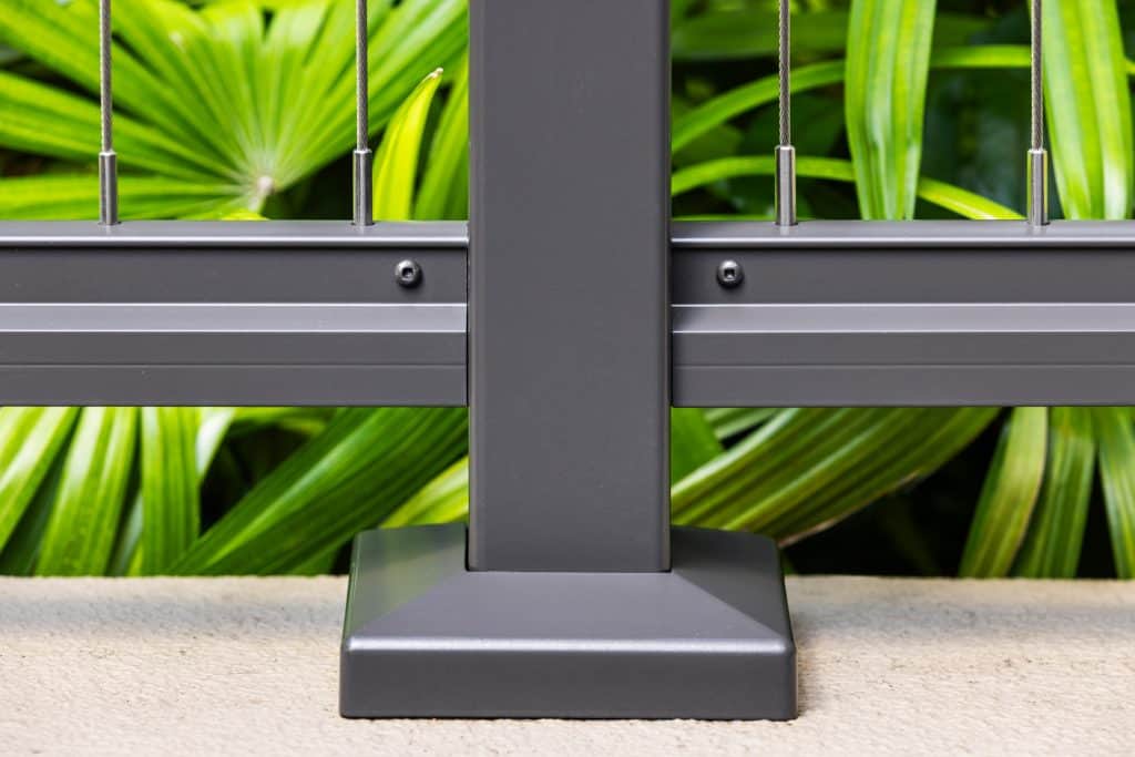 Base detail of a black Feeney cable railing post mounted on a concrete surface at a Honolulu condominium, with lush tropical greenery in the background.